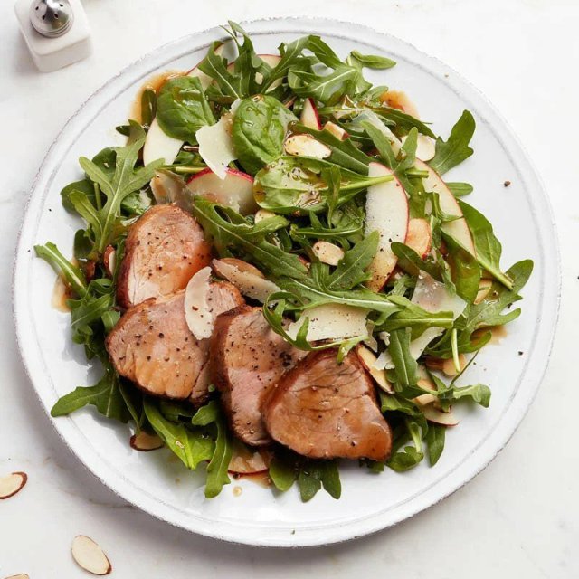 a white plate with green lettuce, slices of pork tenderloin, nuts, shaved parmesan and a brown dressing